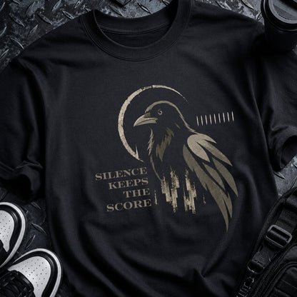 A black short-sleeve graphic T-shirt laid flat on a dark textured surface, featuring a distressed raven illustration and the phrase 'SILENCE KEEPS THE SCORE' in muted beige/gold tones. Partial sneakers and a black bag are visible around the shirt as styling props.