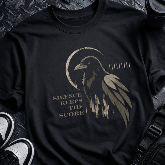 A black short-sleeve graphic T-shirt laid flat on a dark textured surface, featuring a distressed raven illustration and the phrase 'SILENCE KEEPS THE SCORE' in muted beige/gold tones. Partial sneakers and a black bag are visible around the shirt as styling props.